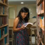 A South Asian woman with glasses reads attentively in a Delhi library, surrounded by bookshelves.